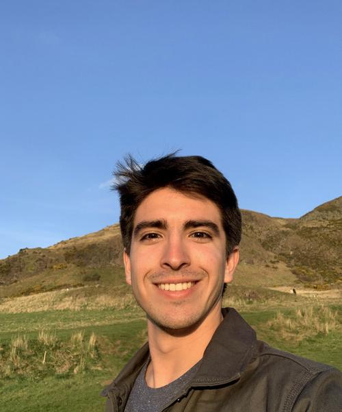 Photo of Ramiro Storni, smiling, wearing a gray t-shirt and brown jacket, with a blue sky and grassy hills in the background